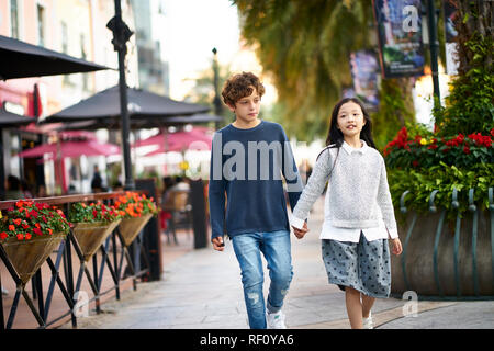 little asian girl and caucasian boy walking together holding hands outdoors Stock Photo