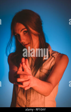 Charming young lady with piercing showing gun gesture and looking at camera in redness on blue background Stock Photo