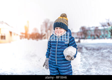A little boy of 3-6 years old is standing outside in winter in the city. In the hands of holding a big snowball. Free space for text. Children's games in the winter. Stock Photo