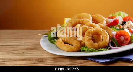 Golden fried calamari rings with fresh Greek salad served on a platter on a wooden counter with copy space in banner format Stock Photo