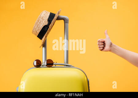 cropped view of woman doing thumb up gesture near travel bag, straw hat and sunglasses isolated on yellow Stock Photo