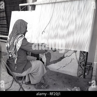 1950s, historical, an arab man in traditional arabian clothing with head scarf sitting on a small chair using a hand loom to weave a decorative rug, Saudi Arabia. Stock Photo