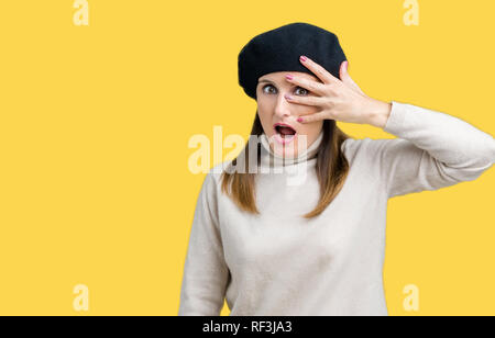 Middle age mature woman wearing winter sweater and beret over isolated background peeking in shock covering face and eyes with hand, looking through f Stock Photo