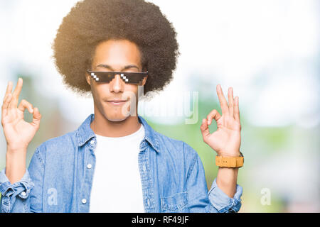 Young african american man with afro hair wearing thug life glasses relax and smiling with eyes closed doing meditation gesture with fingers. Yoga con Stock Photo