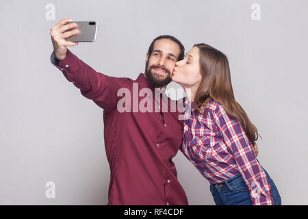 Portrait of happy in love satisfied couple standing and doing selfie, she is kissing her boyfriend and he is smiling at camera of mobile smart phone.  Stock Photo