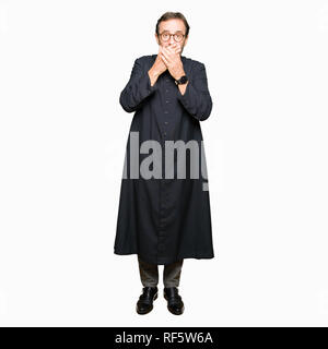 Middle age priest man wearing catholic robe shocked covering mouth with hands for mistake. Secret concept. Stock Photo