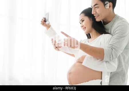 Beautiful Asian pregnant woman and Asian handsome husband holding and looking a sonogram in house. Concept of pregnancy, health care, gynecology, medi Stock Photo