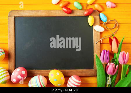 Old blank vintage slate with copy space framed by colorful Easter eggs and fresh spring tulips on a yellow wood background Stock Photo