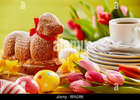 Delicious novelty lamb shaped Easter cake with a red ribbon collar on a spring table set with tulips, Easter eggs, plates and coffee Stock Photo