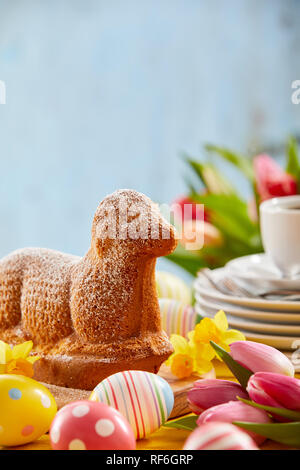 Speciality Easter cake in the form of a lamb on a festive table with colorful patterned eggs, fresh spring daffodils and tulips and a stack of plates  Stock Photo