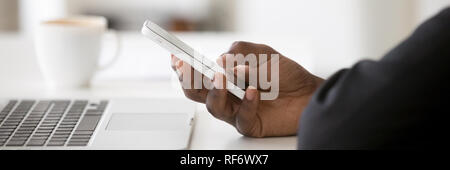 Closeup african employee sitting at desk near laptop holding smartphone Stock Photo