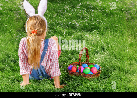 A cute little baby girl in bunny ears is sitting on the lawn, next to a basket of colorful Easter eggs Stock Photo