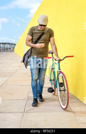 Front view of a young trendy man with a fixed bike wearing casual clothes while using a mobile phone against a yellow wall outdoors in a sunny day Stock Photo