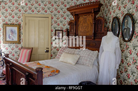 Antique bedroom interior, double bed, ornate wood headboard & footboard, lace pillowcases, wedding dress, vintage floral wall paper, framed portraits. Stock Photo