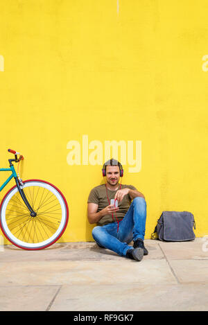 Front view of an attractive man sitting on the floor with trendy look, using a mobile phone having headphones while looking at screen against a yellow Stock Photo