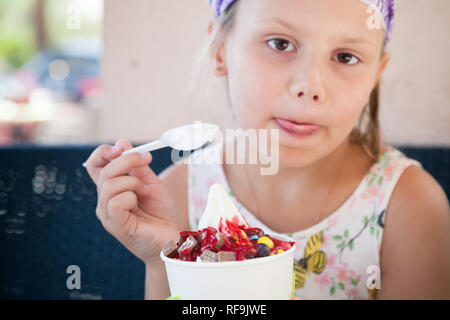 Little blond European girl eats ice cream with fruits, close-up outdoor portrait with selective focus on food Stock Photo