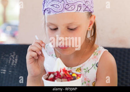 Little blond European girl eats ice cream with fruits, close-up outdoor portrait with selective focus on food Stock Photo