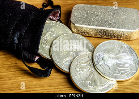 closeup of silver coins american one ounce eagles and brick falling out of black money bag and laying on wooden background Stock Photo