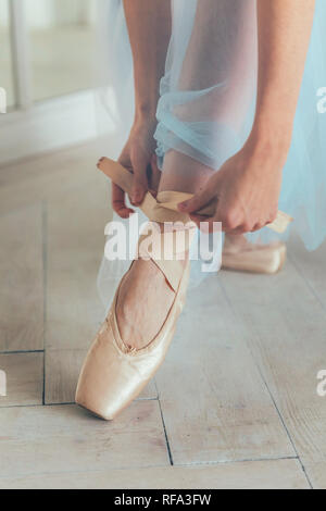 Hands of ballerina in blue tutu skirt puts on pointe shoes on leg in white light hall. Young classical ballet dancer woman in dance class Stock Photo