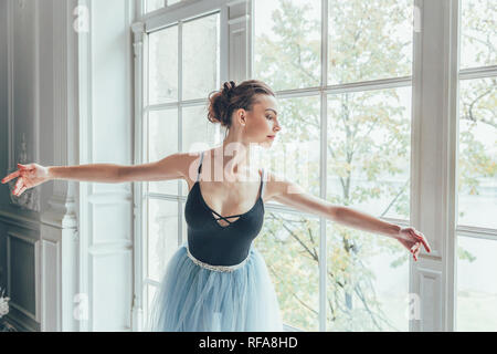 Young classical ballet dancer woman in dance class. Beautiful graceful ballerina practice ballet positions in blue tutu skirt near large window in whi Stock Photo