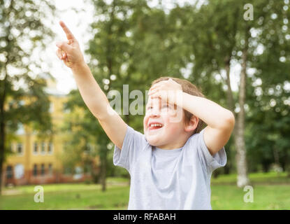 Boy 5 years old looks up at something and shows finger in summer park on background of trees Stock Photo