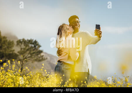 Romantic couple standing outdoors taking selfie.  African man taking selfie on cell phone while woman kissing him on his cheek. Stock Photo