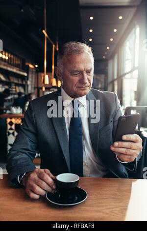 Senior businessman reading text message on his phone while sitting at cafe having coffee. Mature entrepreneur texting on smartphone at coffee shop. Stock Photo