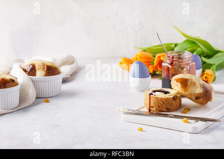 Hot cross buns, butter, jam and colored eggs for Easter breakfast. Flowers in the background. Stock Photo