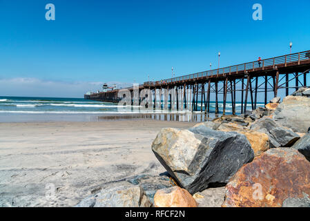 Pier running into sea, large boulders on right with beach and ocean beyond under bright blue sky.