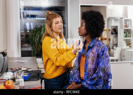 Fair-haired stylist doing makeup to a pretty dark-skinned model Stock Photo