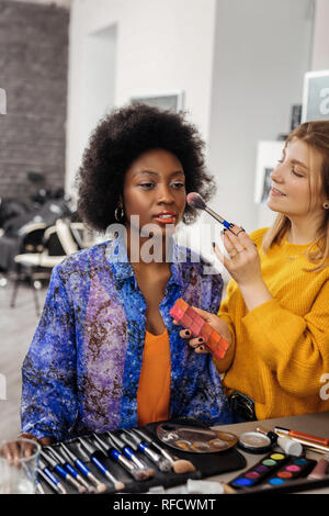 Fair-haired positive stylist doing makeup to the dark-skinned model Stock Photo