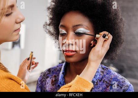 Fair-haired young stylist with black nail polish putting mascara on eyelashes Stock Photo
