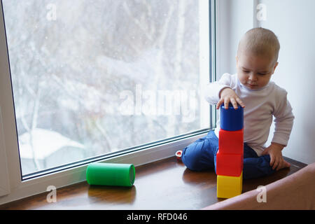 Little boy is sitting on windowsill. He builds a tower of blocks. Winter day outside window. Stock Photo