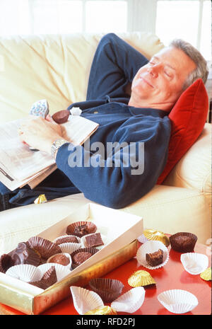 Mature man lounging on sofa with chocolates, TV remote and newspaper, USA Stock Photo