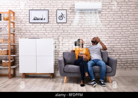 Young African Couple Sitting On Sofa Operating Air Conditioner At Home Stock Photo