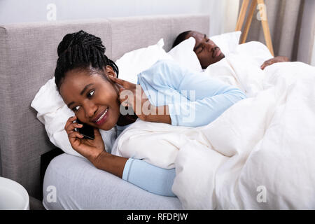 Young African Woman Talking On Cellphone While Her Husband Sleeping On Bed In Bedroom Stock Photo