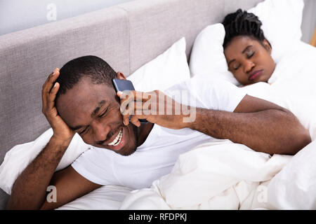 Smiling Young African Man Talking On Smartphone Besides His Wife Sleeping On Bed In Bedroom Stock Photo