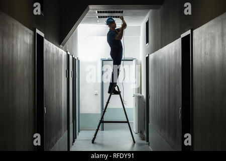 Silhouette Of A Male Electrician On Step Ladder Installing Light At Corridor Stock Photo