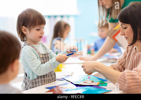 Child with scissors in hands cutting paper with teachers in class room. Group of children doing project in kindergarten. Kids play into team. Stock Photo