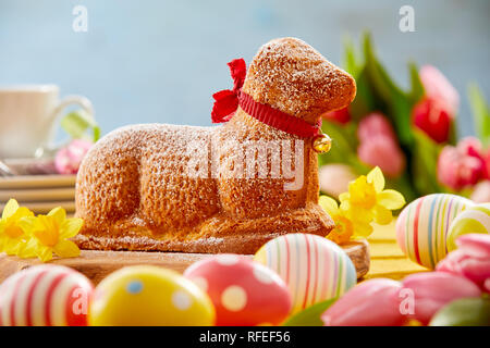 Festive Easter table with a novelty lamb cake tied with a red ribbon surrounded by colored eggs, spring daffodils and tulips Stock Photo