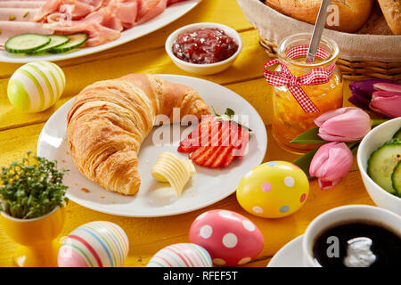 Delicious Easter breakfast on a decorated table with colorful eggs and a freshly baked croissant and strawberry served with jam, cold meats, cheese an Stock Photo