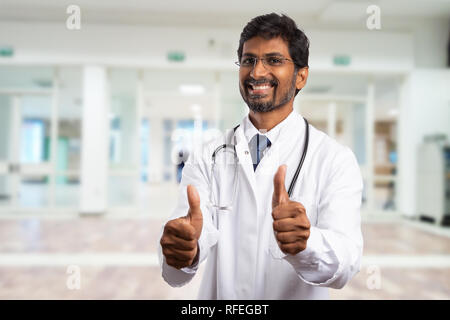 Cheerful indian male medic or doctor making like gesture holding up two thumbs in hospital hallway wearing goggles Stock Photo