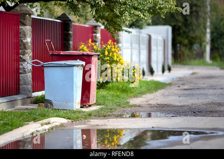 Two steel red and gray painted trash bins of old-fashioned design in backyard along road with puddle as concept of reuse and sorting garbage with copy Stock Photo