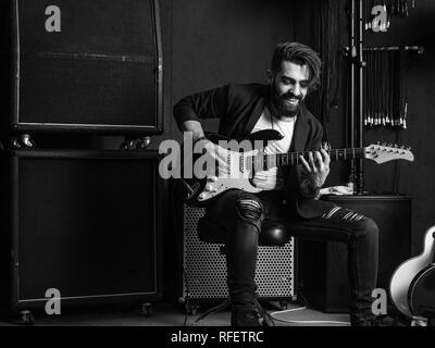 Black and white photo of a man with beard sitting and playing his electric guitar in a recording studio. Stock Photo