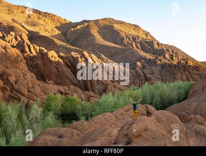 Young man stands on rock and stretches his arms into the air, rock formation Pattes Des Singes, red sandstone rocks