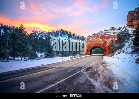 Road with tunnel through red rock arch in snow, at sunset, Highway 12, sandstone rocks, Red Canyon, Panguitch, Utah, USA Stock Photo