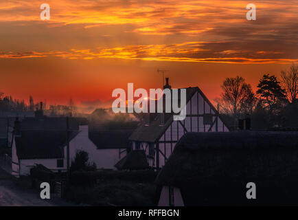sunrise in the village of Monkton, Kent, UK. The sun is just appearing behind a cloud producing a rim light and misty haze enhanced by smoke from a ch Stock Photo