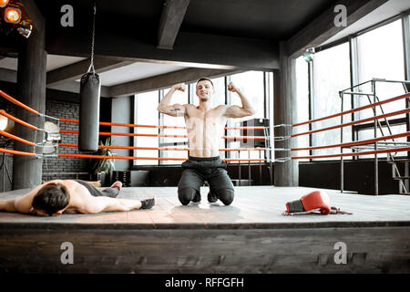 Beaten boxer lying knocked out on the boxing ring with strong man winner showing muscles Stock Photo