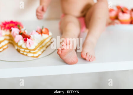 Little feet of baby two years old in pink pants sitting on the white table near the birthday cake and different pink sweets on the table. Stock Photo