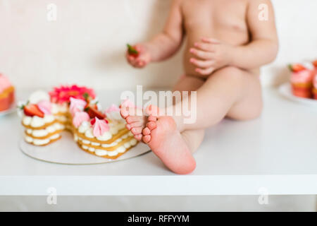 Little blonde baby girl two years old in pink pants sitting on the white table near her birthday cake and different pink sweets on the table. Stock Photo